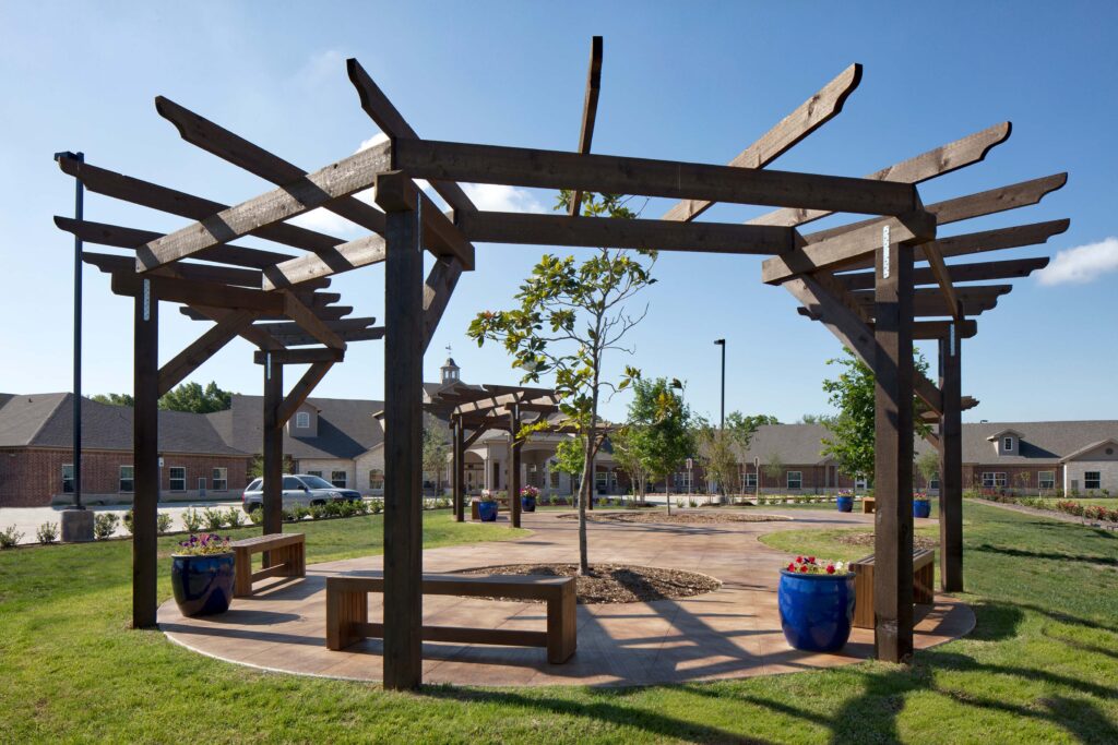 3D3A3882RT1RAB11inch - Twin Rivers A large wooden pergola provides shade over benches in a community courtyard at Twin Rivers, an assisted living and memory care community near Dallas.
