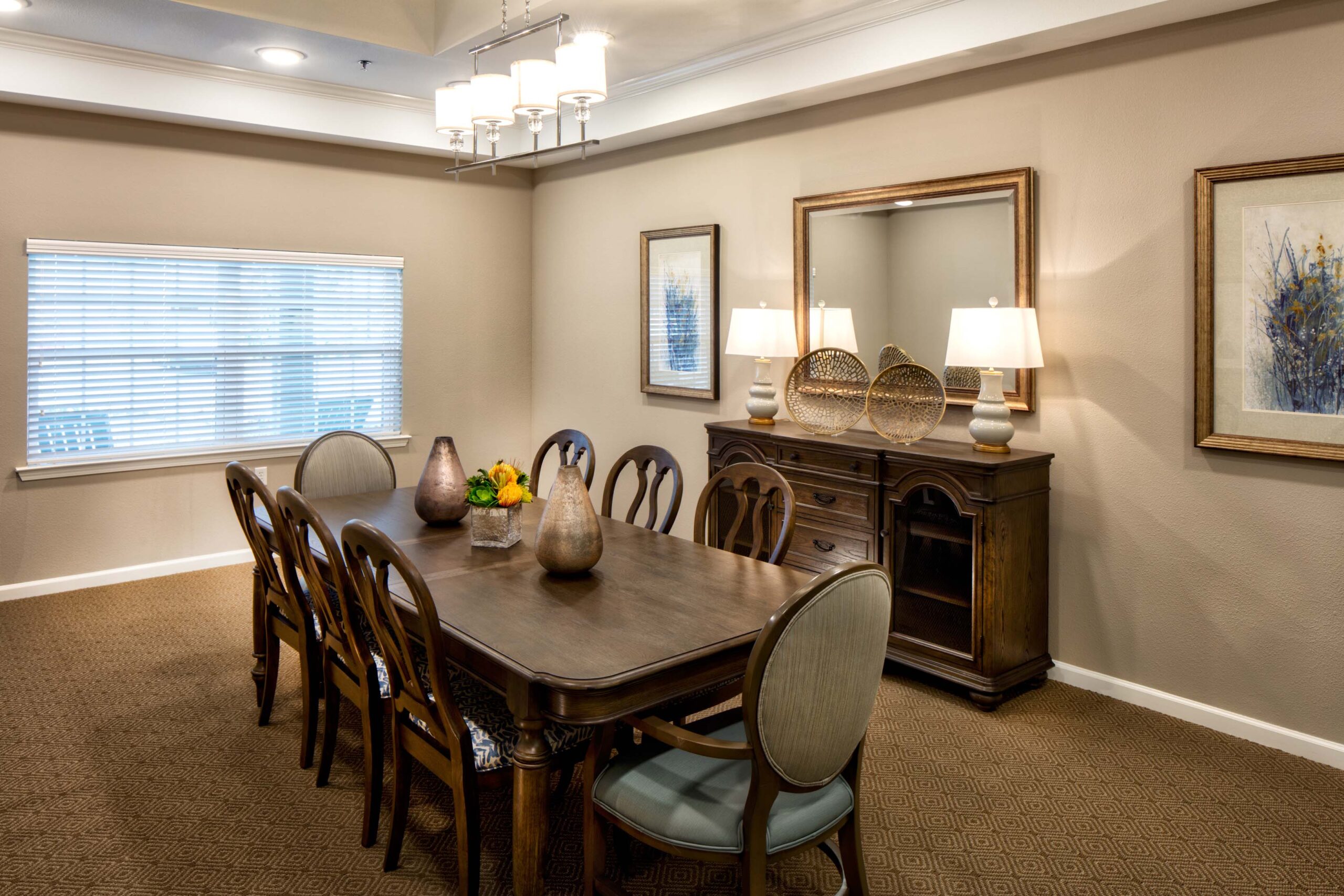 Jim Wilson3D3A5688RT1A - Twin Rivers A long, dark-brown dining table with eight chairs, two table lamps, and a small flower bouquet on the table, a dresser to the right, underneath a mirror, and two lit-up lamps on the dresser at Twin Rivers, an assisted living and memory care community near Plano, TX.